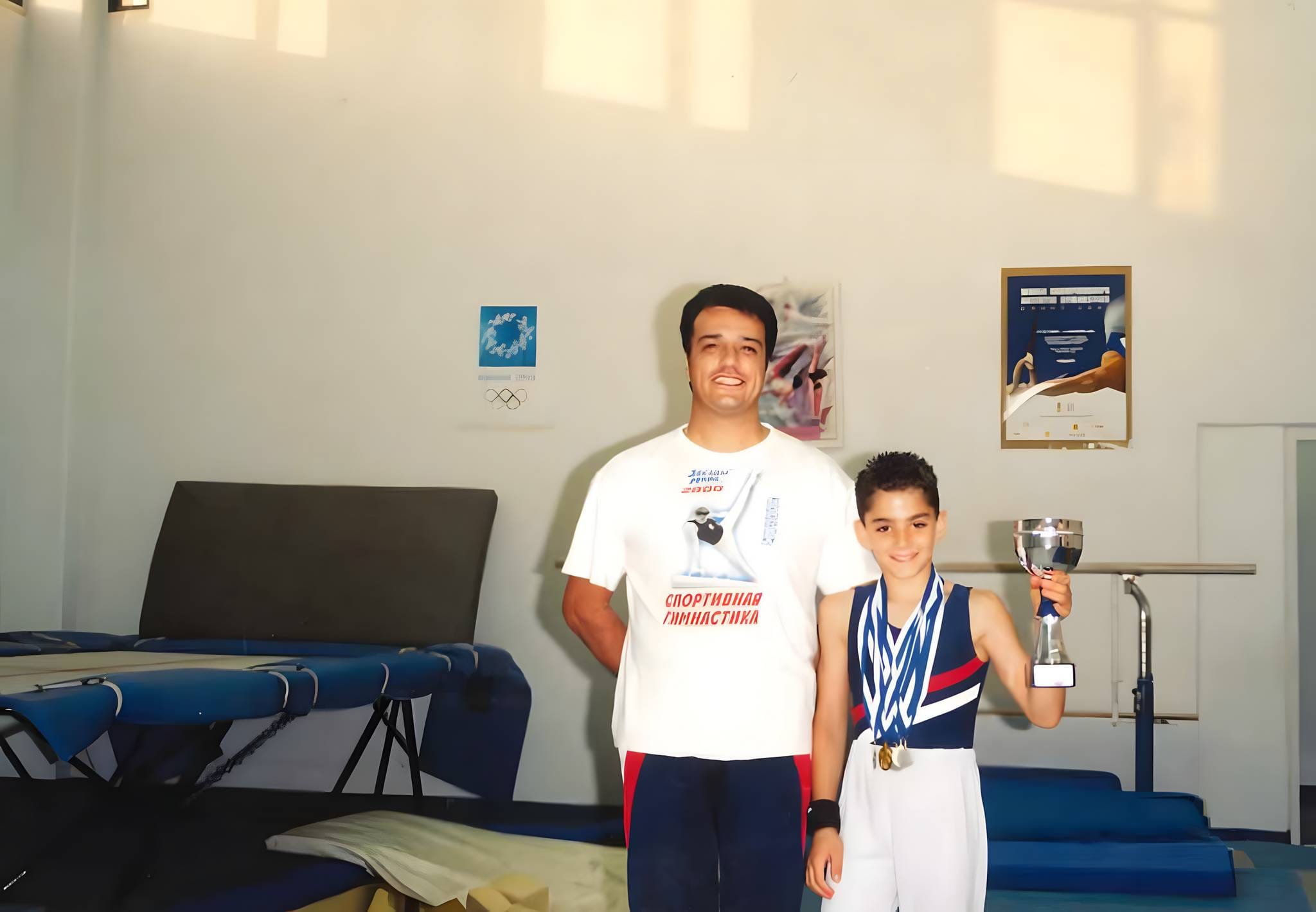 Young Andreas Tricomitis holding his trophy with his coach after a Cyprus national gymnastics title